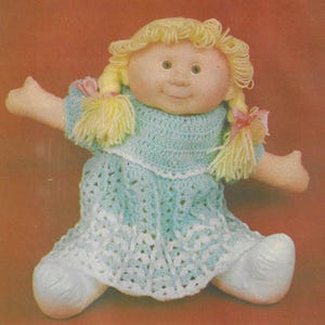 Puede incluir: Un vestido de crochet de color azul claro con ribete blanco. El vestido lo lleva una muñeca con el pelo rubio y una cara sonriente.