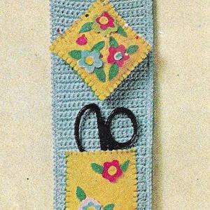May include: A blue crocheted wall organizer with three pockets. The top pocket holds a ruler, the middle pocket holds scissors, and the bottom pocket has a yellow felt panel with colorful flower appliques.