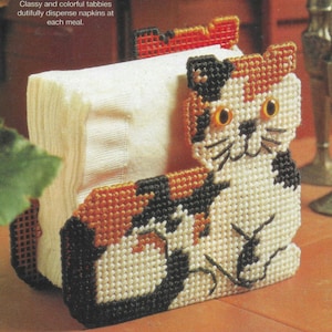 Puede incluir: Un servilletero artesanal con forma de gato calicó, hecho con técnica de punto de cruz. El gato tiene detalles naranjas, negros y blancos, con ojos amarillos y bigotes negros. El soporte dispensa servilletas blancas. El texto dice: Classy and colorful tabbies dutifully dispense napkins at each meal.