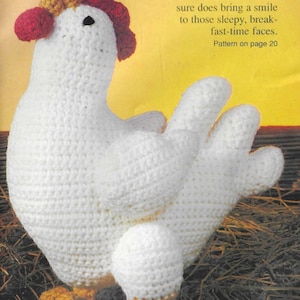 Puede incluir: Un gallo de crochet blanco con detalles rojos y amarillos se sienta en un lecho de paja. El gallo tiene un huevo blanco debajo de su cuerpo. El texto "Rise 'n Shine" está escrito sobre el gallo.