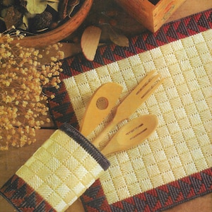 May include: A set of two white plastic canvas placemats with a red, black, and white geometric pattern border. The placemats are decorated with wooden spoons and a wooden utensil holder.