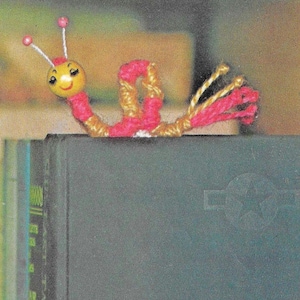 May include: A yellow and pink yarn snail bookmark with a smiling face. The snail is perched on the edge of a book.