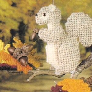 May include: A crocheted squirrel ornament with a brown tail and a white body. The squirrel is holding a brown acorn in its paw. The ornament is surrounded by crocheted leaves and acorns in shades of yellow, brown, and orange.