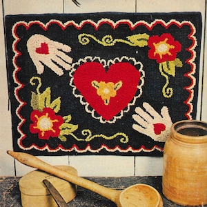 Może przedstawiać: Ręcznie robiony dywan z wzorem serca w środku, otoczony dwoma dłońmi i dwoma kwiatami. Dywan jest wykonany z wełny i ma ciemnoniebieskie tło.