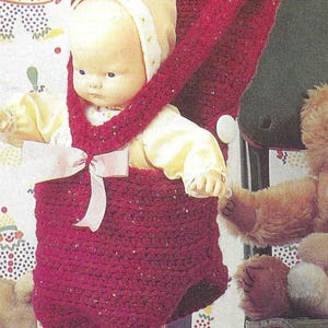 May include: A red crocheted baby carrier with a pink ribbon and white buttons. The carrier is hanging from a metal bar and a doll is inside.