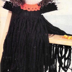 Può includere: Una bambola di strega all'uncinetto con un cappello nero, bordi arancioni e neri e un vestito nero. La bambola ha un viso bianco con occhi neri e un sorriso rosso. I capelli della bambola sono marroni e irsuti. La bambola è appesa a uno scaffale di legno.