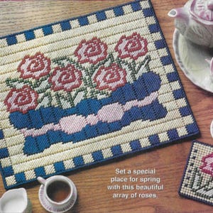May include: A needlepoint placemat with a floral design featuring red roses, blue accents, and a cream background. A matching coaster and a tea set are also visible. The text reads: "Set a special place for spring with this beautiful array of roses."