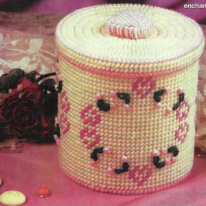 May include: A white crocheted canister with a pink and black floral design. The canister has a lid with a pink and white crocheted top.