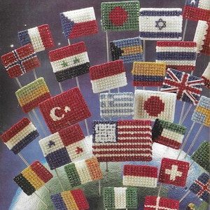 Könnte beinhalten: Eine Sammlung bunter, mit Kreuzstich bestickter Flaggen aus aller Welt, darunter die Vereinigten Staaten, Kanada, Frankreich, Deutschland und viele mehr. Die Flaggen sind auf kleinen Holzstäbchen montiert und in einem Kreis auf einem Globus angeordnet.