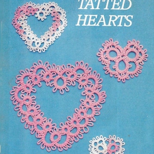 Puede incluir: Cuatro corazones tejidos en rosa y blanco. Los corazones son de diferentes tamaños y tienen un diseño delicado y de encaje. El título "TATTED HEARTS" está impreso en negro en la parte superior de la imagen.