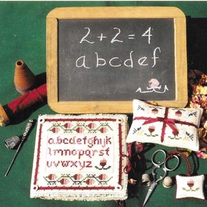 Puede incluir: Un conjunto de tres proyectos de bordado de punto de aguja que incluyen una pizarra con la ecuación "2+2=4" y las letras "abcdef", un cuadrado con el alfabeto y una pequeña almohada con un diseño de flor roja.