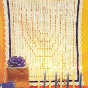 May include: A white crocheted menorah wall hanging with a blue border and a scalloped edge. The menorah is decorated with a traditional menorah design. The menorah is hanging on a wall with a yellow background. A menorah with lit candles is in the foreground.