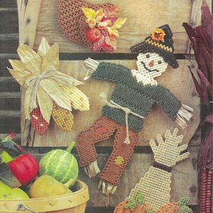 May include: A collection of autumn-themed plastic canvas crafts, including a scarecrow, a cornucopia, a pumpkin, and a bundle of corn. The crafts are displayed on a wooden background.