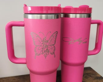 May include: Two bright pink stainless steel tumblers with handles and lids. The left tumbler features a butterfly graphic with the text Love Like Dolly. The right tumbler displays the signature Dolly xoxo in a script font. Both have a textured finish.