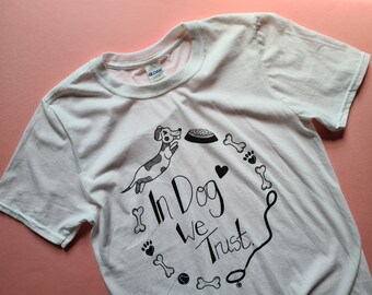 May include: White t-shirt featuring a black and white illustration of a dog, a bone, a heart, and a bowl. The text "In Dog We Trust" is written in black.