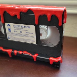 May include: A black VHS tape with a red, dripping substance on the top and bottom. The tape is labeled "Sleepy Hollow Special Edition" with the words "Tim Burton's" and "Mini-Series" printed below.