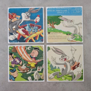 May include: Set of four square ceramic coasters featuring illustrations from classic Warner Brothers cartoons. The coasters feature Bugs Bunny, Porky Pig, and Sylvester the Cat. The coasters are printed with a blue and white background and feature the text "A Whitman Book" and "Warner Brothers Cartoons".