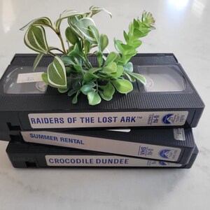 May include: A stack of three black VHS tapes with labels reading "Raiders of the Lost Ark", "Summer Rental", and "Crocodile Dundee". Green and white leafy plant decorates the top. The tapes are on a white surface.