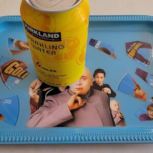 May include: A rectangular, light blue tray with a beaded edge, featuring a collage of images and a yellow can of Kirkland Sparkling Water. The can is labeled "lemon" and is 12 fl oz. The tray's design includes images of characters.