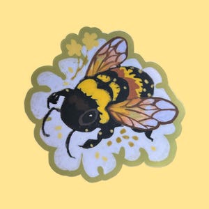 May include: A die-cut sticker featuring a bumblebee with black and yellow stripes, a fuzzy black head, and translucent wings, resting on a white flower with yellow centers and small yellow accents, outlined in a thin gold border.