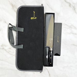 May include: A chef's knife with a black handle and silver blade, presented in a black box and carrying case. The knife is labeled with the brand name "HANAKO" in gold lettering. The carrying case is black with a gray trim.