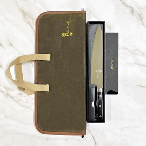 May include: A chef's knife with a black handle and silver blade, presented in a black box. The knife is accompanied by a sharpening stone and a brown canvas knife roll with a tan handle. The brand name "HANAKO" is visible.
