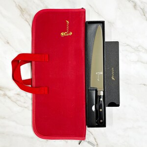 May include: A chef's knife with a black handle and silver blade, presented in a black box. The box is next to a red knife case with a handle. The knife and case have the brand name "HANAVA" printed on them.