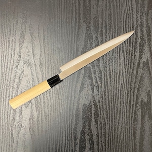 May include: A long, silver kitchen knife with a wooden handle. The knife is lying on a dark wood surface.