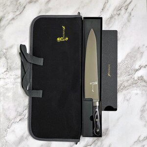 May include: A stainless steel chef's knife with a black and gray handle is displayed in a black box. The knife is next to a black carrying case with the word "HANAKO" in gold lettering. The knife is designed for professional use.