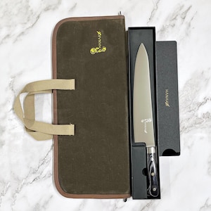 May include: A chef's knife with a silver blade and a black and gray handle, presented in a black box. The knife is displayed next to a brown canvas knife roll with a tan handle and the word "HANAKO" printed on the box and roll.