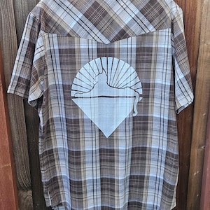 May include: Short-sleeved button-down shirt with a brown, tan, and blue plaid pattern. The back features a white graphic of a cat silhouette within a sunburst design and a diamond shape. The shirt is hanging on a black hanger.