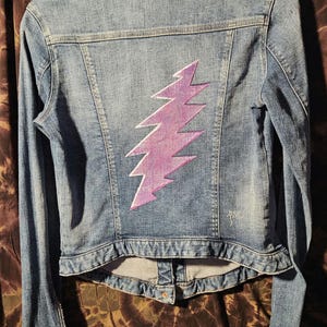 May include: Light blue denim jacket with a purple and white lightning bolt design on the back. The jacket has long sleeves, a button closure, and classic denim jacket details.