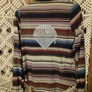 May include: A long-sleeve button-down shirt featuring horizontal stripes in shades of navy blue, maroon, light blue, and grey. The back of the shirt displays a white graphic print of a cat silhouette resting atop a diamond shape with sun rays.