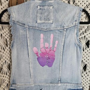 May include: A light-wash blue denim vest featuring a sleeveless design with frayed armholes, a classic collar, and a central back graphic of a hand making a sign language gesture in a gradient of pink to purple colors on the back panel.