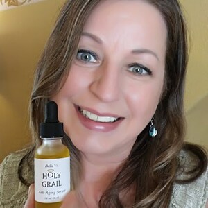 Puede incluir: Una mujer sostiene una peque&ntilde;a botella de vidrio &aacute;mbar con un gotero. La etiqueta de la botella dice "Bella Ve Holy Grail Anti-Aging Serum 1 fl oz (30 ml)".