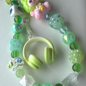 May include: A beaded keychain featuring a variety of green and clear beads, including a light green headphone charm and a cartoon character. The keychain also includes a star-shaped bead with the word "STAR" printed on it. The beads are various shapes and sizes.