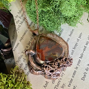 Puede incluir: Un collar con colgante de cobre hecho a mano. El colgante presenta una piedra en forma de lágrima con patrones rojos, marrones y crema, engastada en un marco de cobre texturizado. El collar se exhibe sobre una página de libro vintage.