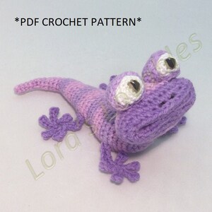 May include: A crocheted purple and pink lizard with white eyes. The lizard has a long tail and four legs with three toes each. The lizard is lying on its side with its head turned to the right. *PDF CROCHET PATTERN*