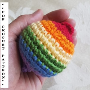 May include: A rainbow crocheted ball with red, orange, yellow, green, blue and purple stripes. The ball is being held in a hand.