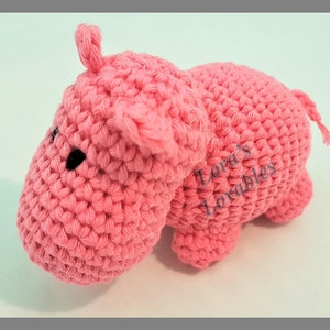 May include: A pink crocheted hippopotamus toy. The toy has black eyes and a small tail. The toy is made by Lora's Lovables.