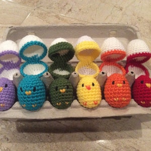 May include: A set of eight crocheted chicks in an egg carton. The chicks are in various colours, including purple, teal, green, yellow, orange, and red. Each chick is sitting in a crocheted egg shell.