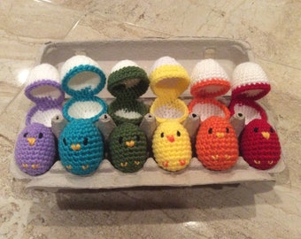 May include: A set of eight crocheted chicks in an egg carton. The chicks are in various colors, including purple, teal, green, yellow, orange, and red. Each chick is sitting in a crocheted egg shell.