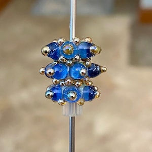 May include: A cylindrical glass bead featuring a white core with three horizontal rows of translucent blue glass protrusions. The bead is accented with small, reflective gold-toned metallic spheres embedded between the blue glass elements.