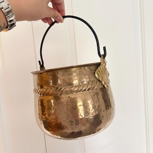May include: A hammered copper bucket featuring a rounded body with a decorative rope-like band around the upper section. The bucket includes a black metal swing handle attached to ornate brass-coloured side mounts with a shell-like design.