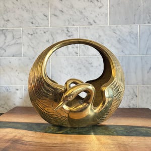 May include: A gold-coloured brass sculpture of two swans intertwined, forming a circular basket shape. The sculpture has a handle and detailed feather textures. The piece is likely a decorative item.