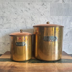 May include: Two cylindrical brass canisters with matching lids. The smaller canister features a silver-toned label reading COFFEE, while the larger canister features a silver-toned label reading FLOUR. Both containers have a polished metallic finish.
