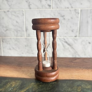 May include: A wooden hourglass featuring a medium brown finish with a smooth, rounded top and base connected by four turned, spindle-style pillars. A clear glass chamber is positioned in the centre, containing fine white sand.