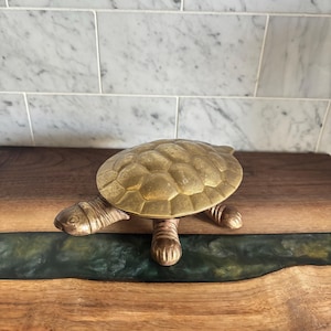 May include: A brass turtle ornament with a textured shell and detailed legs and head. The turtle is on a wooden surface with a dark green epoxy river. The background features white marble tiles.