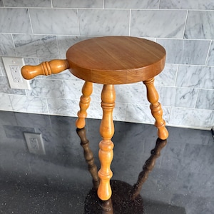 May include: A small wooden stool with a round seat and three turned legs. It features a handle on one side. The wood has a warm, natural finish. The stool is likely used for seating or as a decorative item.