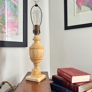 May include: A table lamp with a carved, light beige stone base featuring a classic urn-shaped body and a square pedestal. The lamp includes a brass socket, a metal harp, a silver-toned finial ring, and a clear glass Edison-style light bulb.